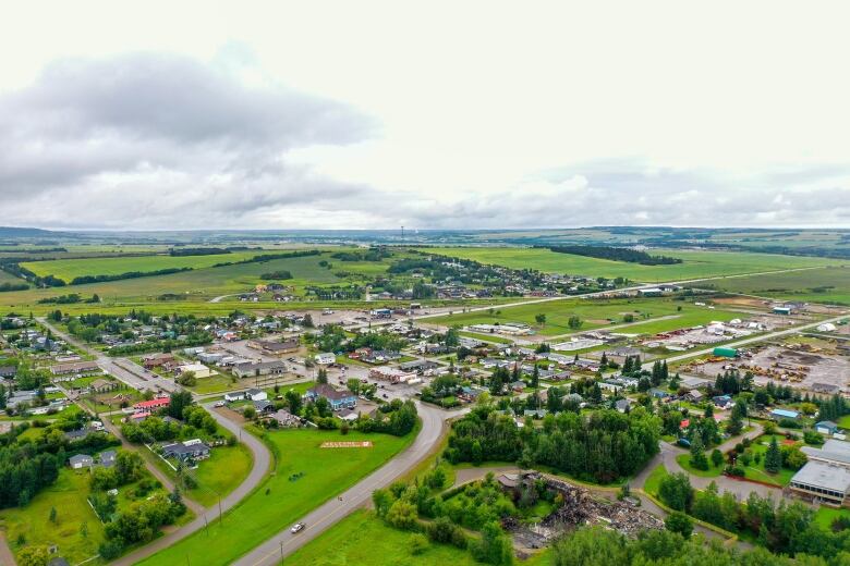 An aerial shot shows a small town surrounded by green fields and trees below a grey, cloudy sky.