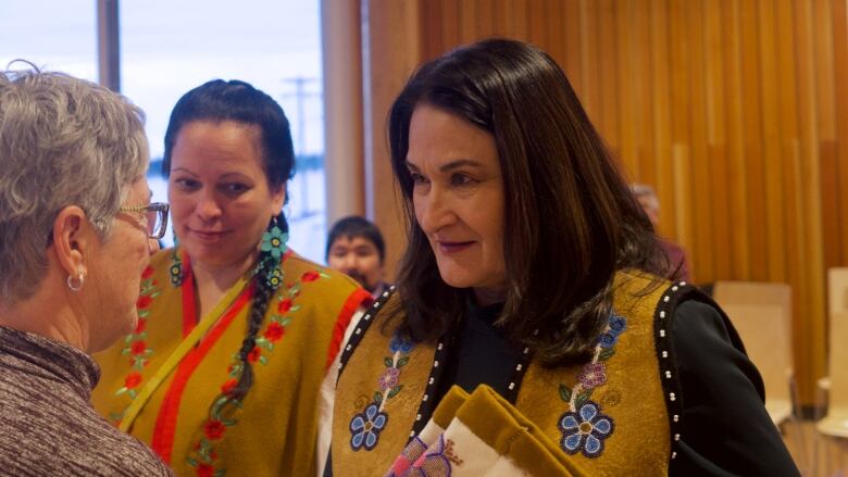 Two women in beaded hide vests stand side-by-side as another woman faces them, inside a room with people in the background.