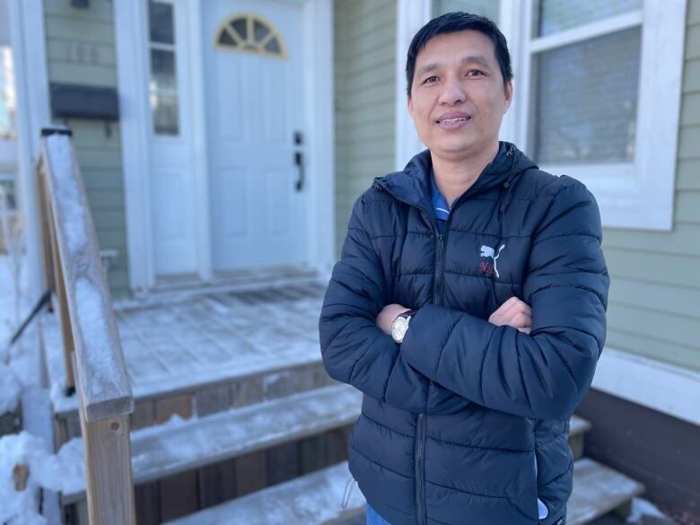 A man with folded arms stands near the steps and front door of a home. 