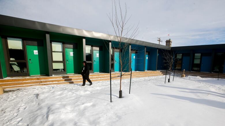 A woman walks through snow next to a black building fronted by blue and green doors next to windows.