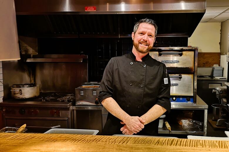 Chef Mathias stands in front of his restaurants kitchen with his hands crossed in front. He is wearing a black chef's jacket.