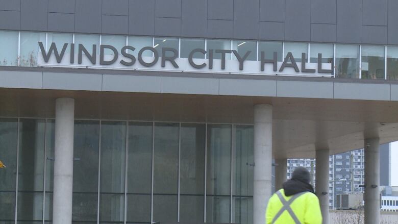 A building with a sign marking it Windsor's city hall with a person wearing a high visibility jacket walking by. 