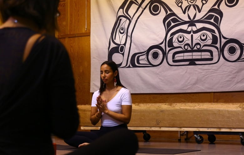 A female yoga teacher sits, legs crossed, with her hands in a prayer position and her eyes closed, in front of a class. 