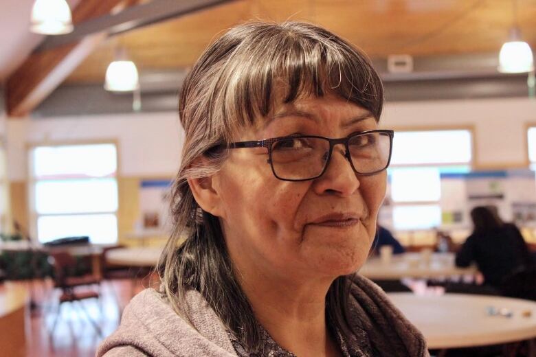 A woman with long dark hair, pulled into a half pony tail, wearing black glasses, looks into the camera.