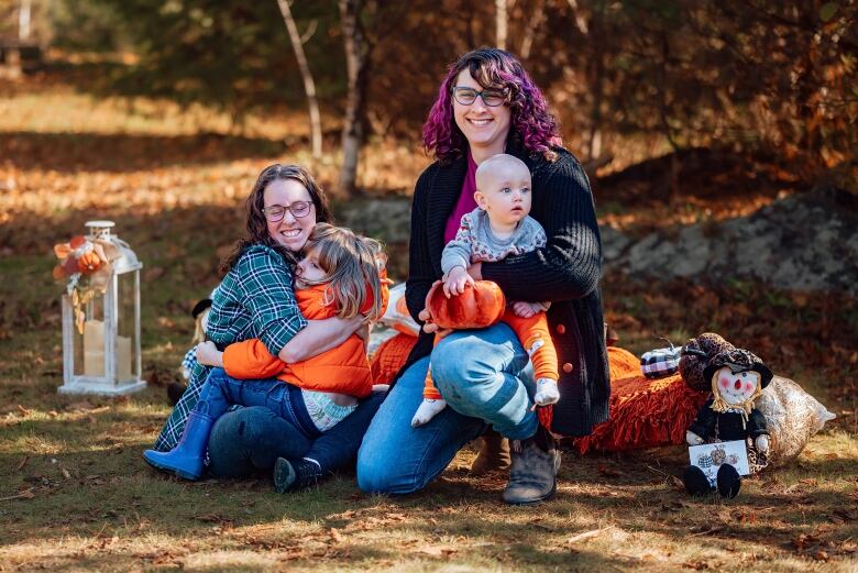 Two adults are outside on the grass each holding a child. The child on the right is a toddler and the other child is older. Both children are wearing orange and the background is autumnal.