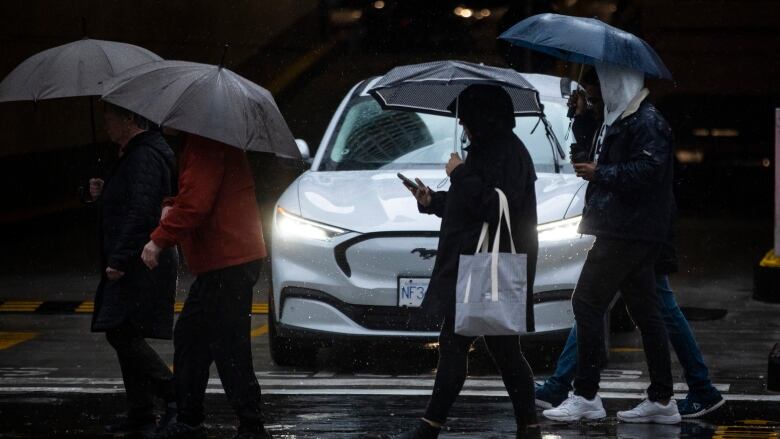 Four people with umbrellas walk in the rain on a street in front of a white car in the dark.