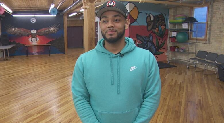 A man in a turquoise hoodie faces the camera. Behind him are paintings of a buffalo, eagle and medicine wheel on the wall.