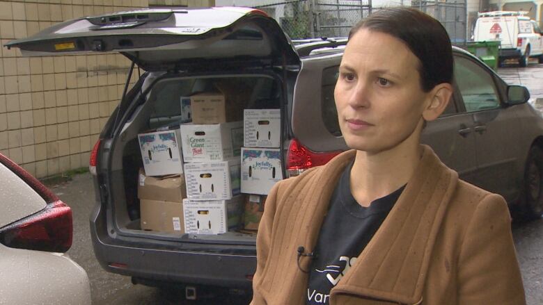 A van door is open with boxes full of food in the back. A woman wearing a black t-shirt, and brown jacket with straight black hair looks at the camera.