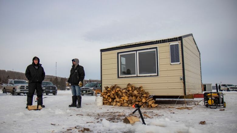 Two men stand outside an ice fishing shack.