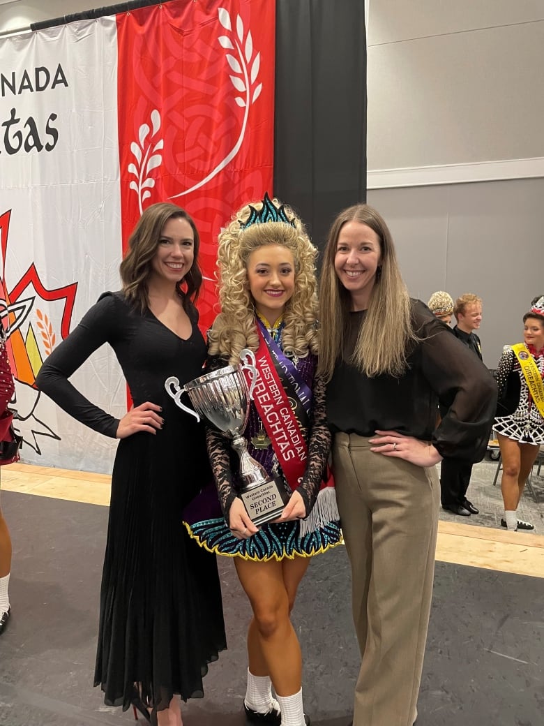 Three women pose for a picture. The one in the centre has an elaborate blonde wig and is carrying a trophy.