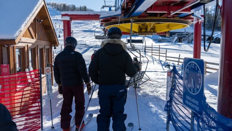 Two skiers are seen from behind, preparing to board a chairlift at a ski hill.