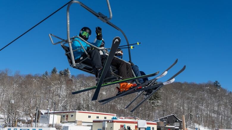 A downhill skier waves at the camera from a chairlift on a sunny winter day.