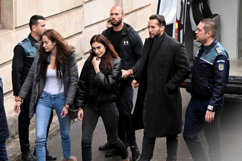 Two women handcuffed to each other are followed by two men handcuffed to each other, surrounded by police as they walk into a building.