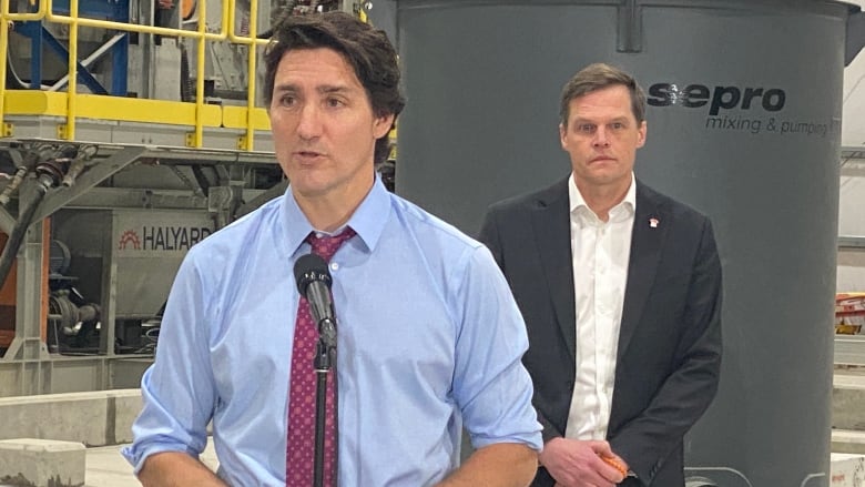 Two white men are standing in a factory. One is standing in front of a microphone. He has long dark hair, and wearing a light blue dress shirt with the sleeves rolled up to his elbow, and purple patterned-tie. The second man, who has dirty blonde hair parted to the left, is standing in the background. He is wearing a black blazer and white button-up shirt. He's wearing a pin on his lapel.