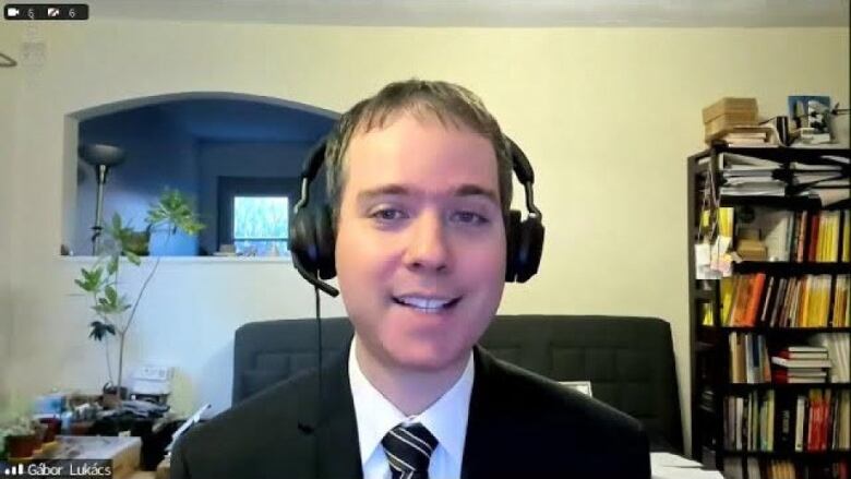 A man wearing headphones and a suit smiles on a Zoom meeting. 