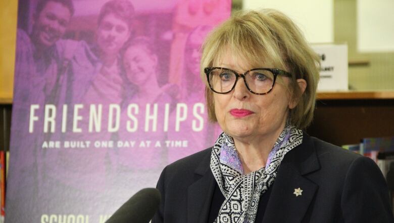 A woman stands by a microphone with a pink poster in the background.