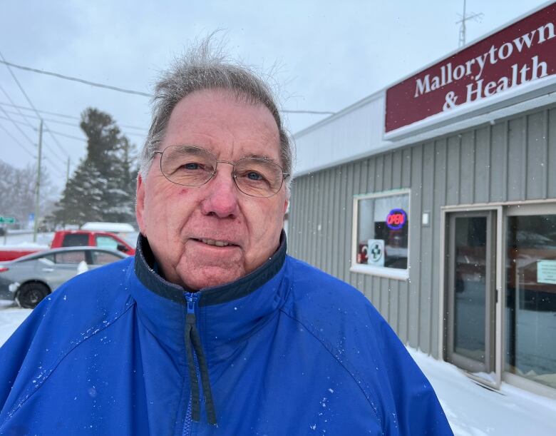 An older man with glasses and wearing a blue winter coat stands in the snow in front of a pharmacy.