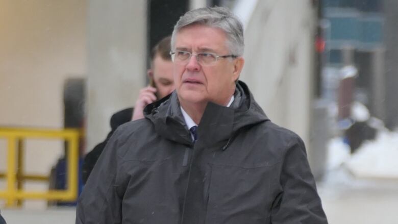 A man with grey hair and glasses, wearing a parka and gloves and carrying a leather bag, walks along a snowy sidewalk.