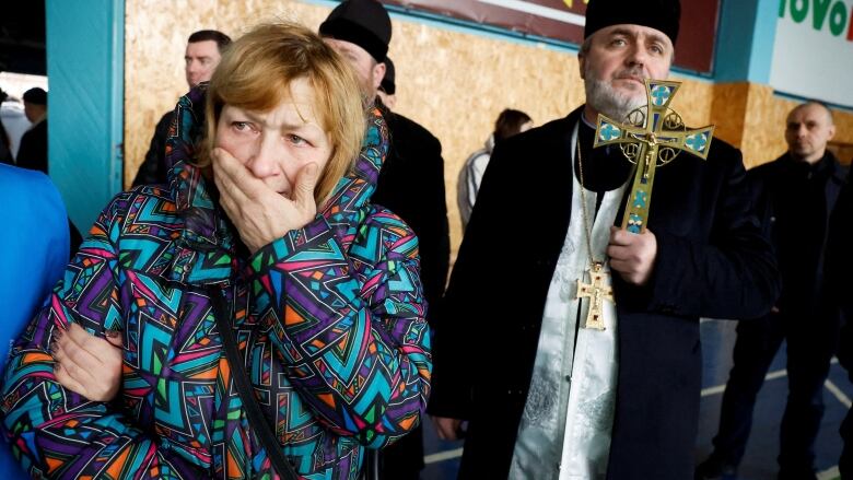 A woman stands with her hand to her mouth in grief, beside a man holding a cross and wearing religious garments.