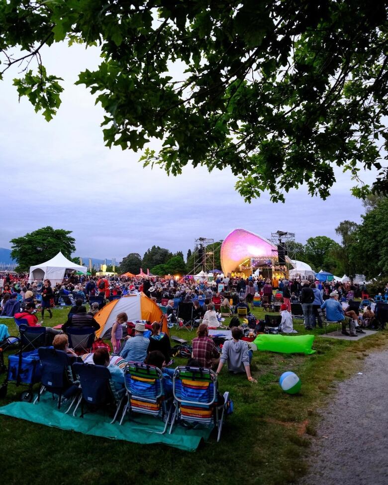 Concert goers sit on the grass at dusk in the summer. A stage with musicians is pictured in the background.