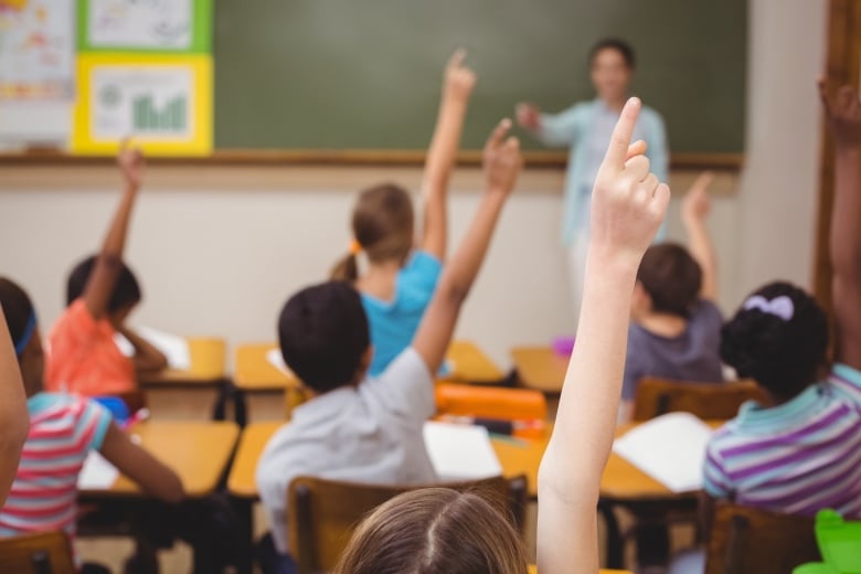 Blurred teacher teaching a lesson in class at the elementary school. Students have their hands up as you look at the back of their heads.