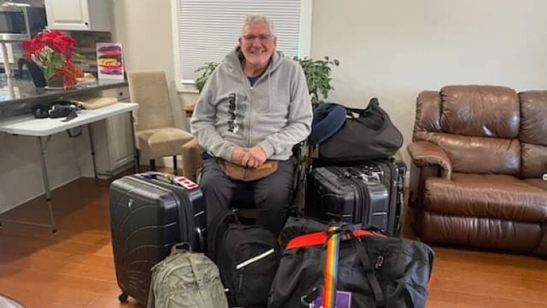 Jim Hamilton, a 63-year-old man in glasses, smiles at the camera. He's sitting in a wheelchair surrounded by bags and suitcases.