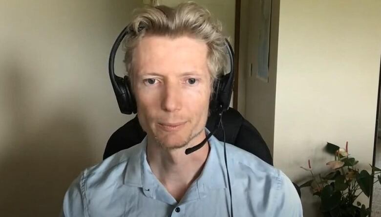 A man with blonde hair and a blue shirt sits at a desk wearing a head set. 