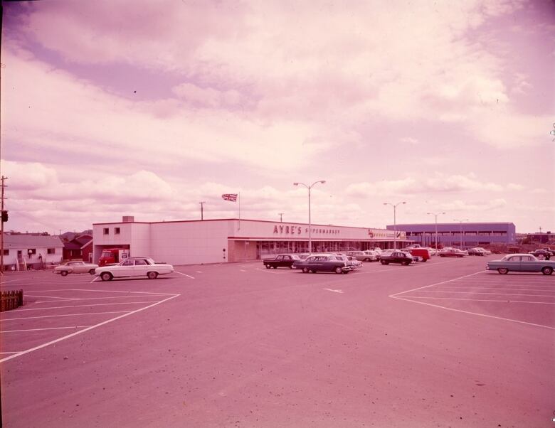 An aged photograph of a white building with a large parking lot in front. The sign on the building reads Ayres Supermarket.