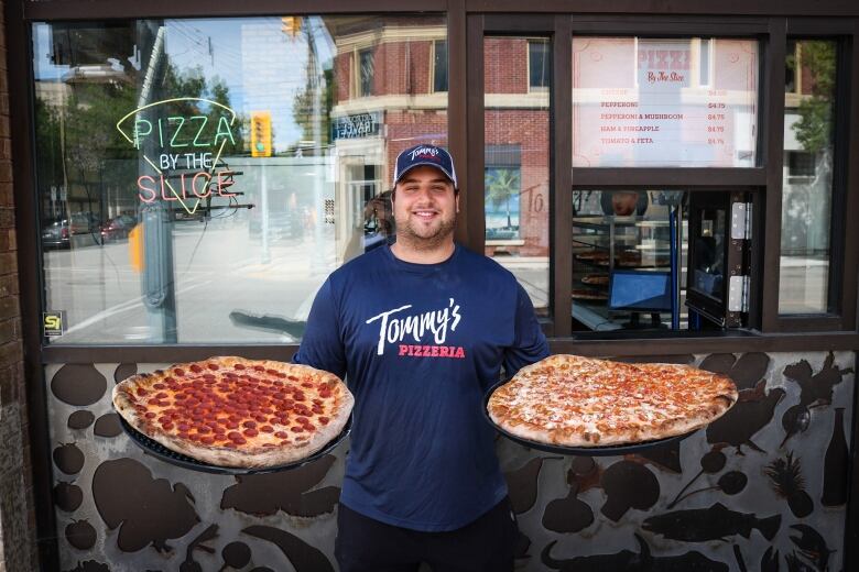 A man stands outside his business and holds a pizza in each hand.