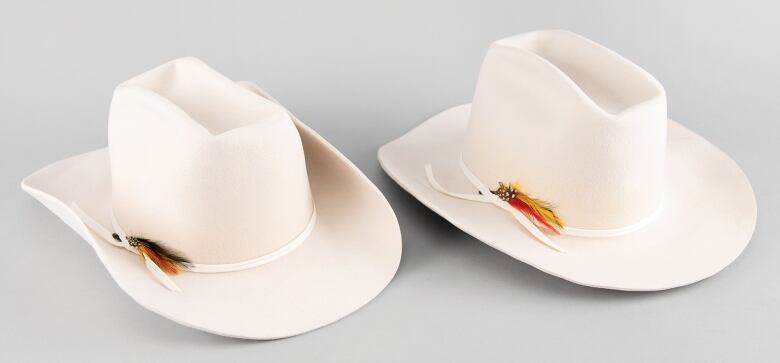 Two white cowboy hats, one with the rim folded, and each with a  white band and feathers. 