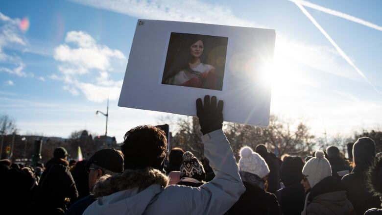 A white poster bearing the image of a young woman is held up by a protester standing among others at a rally.