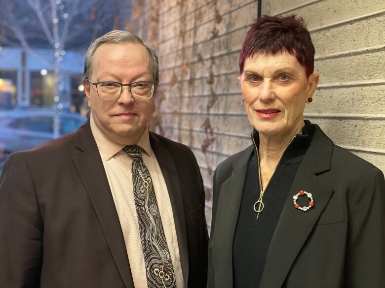 A man and woman stand in suits against a concrete wall.