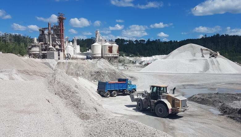 Various machines work in piles of sand and rock, with a lime kiln in the background, underneath a blue sky 