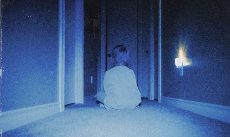 Still frame from the film Skinamarink. Grainy, blue-tinted image of a boy sitting in a hallway.