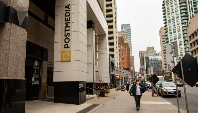 A vertical sign that reads Postmedia is attached to a column outside a downtown building on a busy street as a man carrying a backpack strolls by.