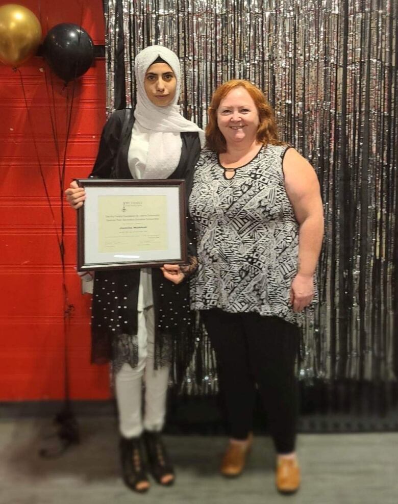 A woman holds a plaque while standing next to another woman in a black and white shirt in front of balloons and streamers on the wall behind them. 