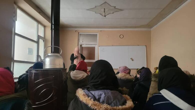 A teacher stands in front of a classroom of girls wearing winter jackets. Her face is blurred.