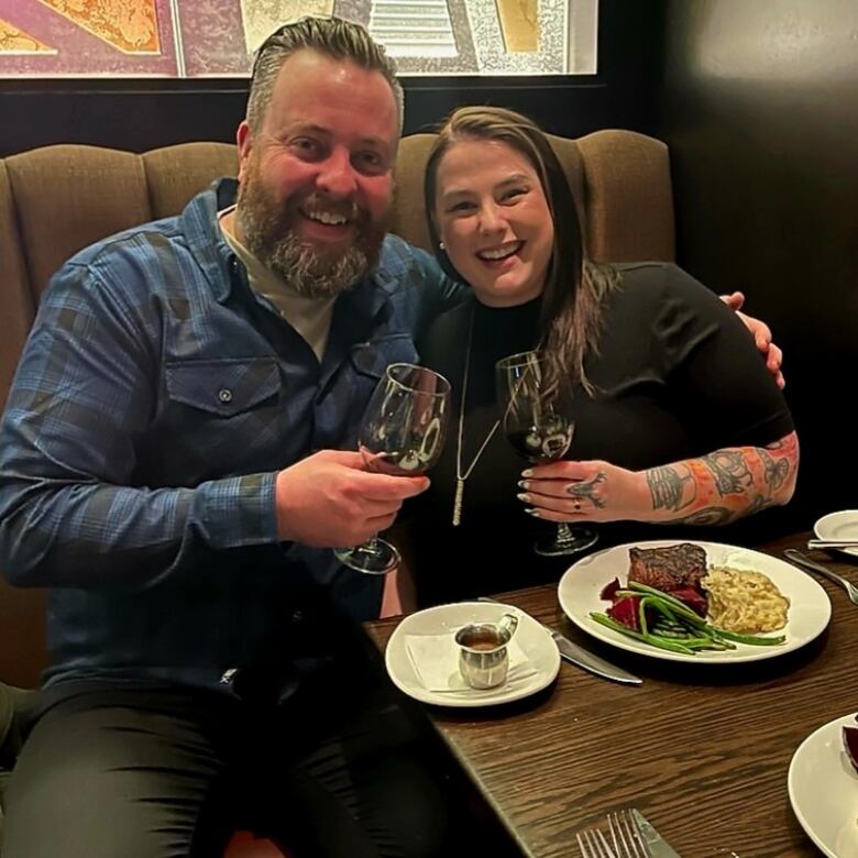 A bearded man sits at a restaurant table next to a woman with tattoos on her arm. The two are holding wine glasses with plates in front of them.