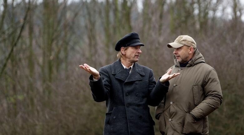 Two men in overcoats and cabs stand side-by-side in a field during the filming of a movie. 