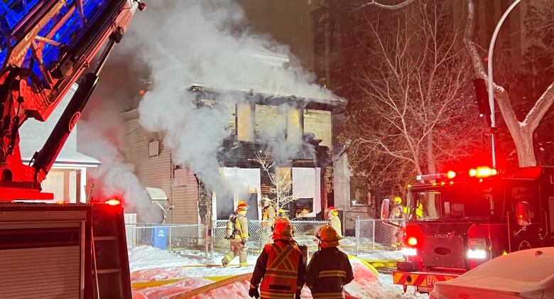 Smoke pours out of a two-storey house in the centre of the photo. Firefighters stand in the foreground wearing helmets and protective coats. Much of the photograph is highlighted in red from the emergency lights on fire trucks.