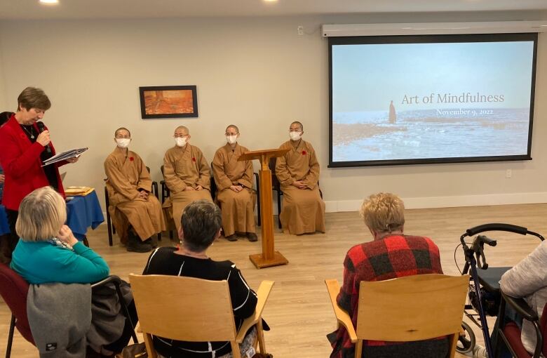 People seated at desks watch a Powerpoint presentation, with four Buddhist nuns seated beside the screen.