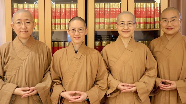 Four Buddhist nuns in beige robes smile for the camera, standing in front of bookshelves lined with thick books.