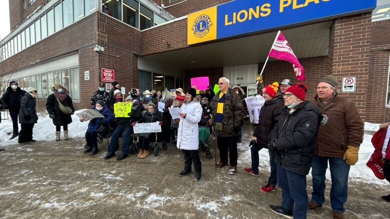 A crowd of people, some holding colourful signs, stand in front of a building with the name Lions Place in yellow letters on a blue background.