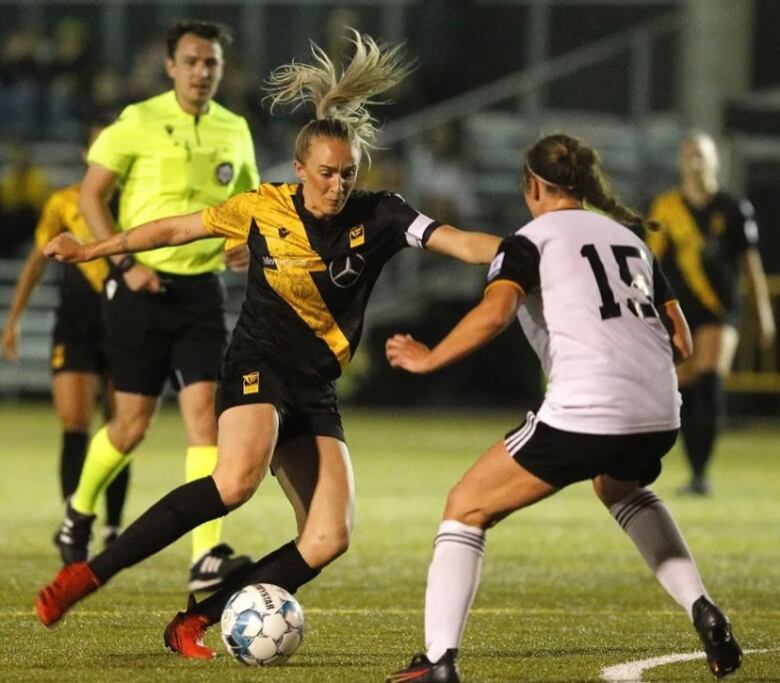 Two women on a soccer field kick a soccer ball. 