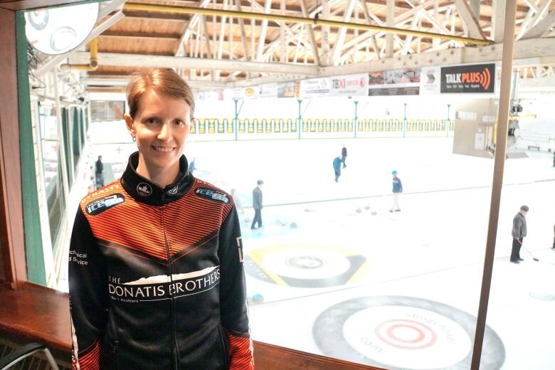 A woman stands next to a window overlooking a curling rink.