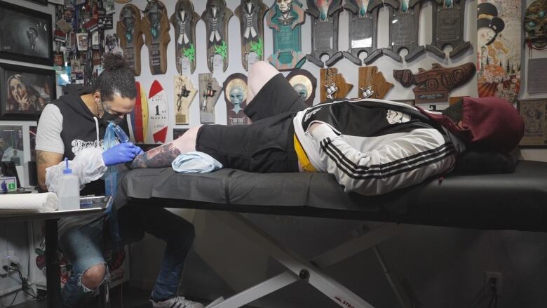 A man lies on a table as another man tattoos his calf.