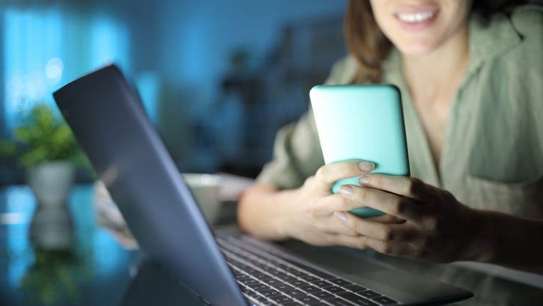 A woman looks at a cellphone while she leans on a desk with a laptop.