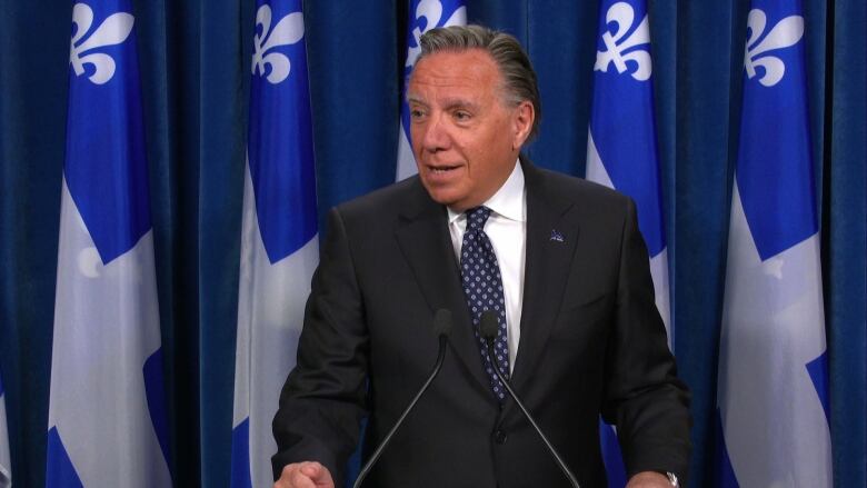 A man in a black suit stands at a podium with blue flags behind him. 