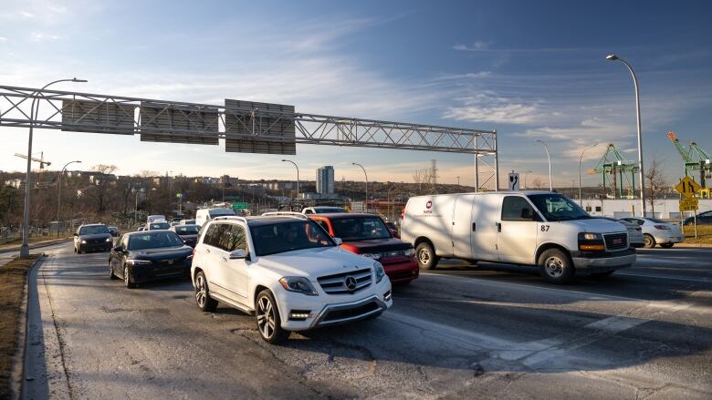 Multiple cars and trucks sit in three lanes of traffic while waiting for a light. Green cranes are visible to the right in the nearby container terminal.