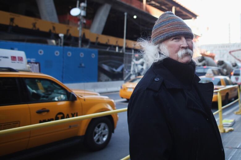 A man with white hair, wearing a hat and coat, is pictured on a street as taxis pass by in the background.